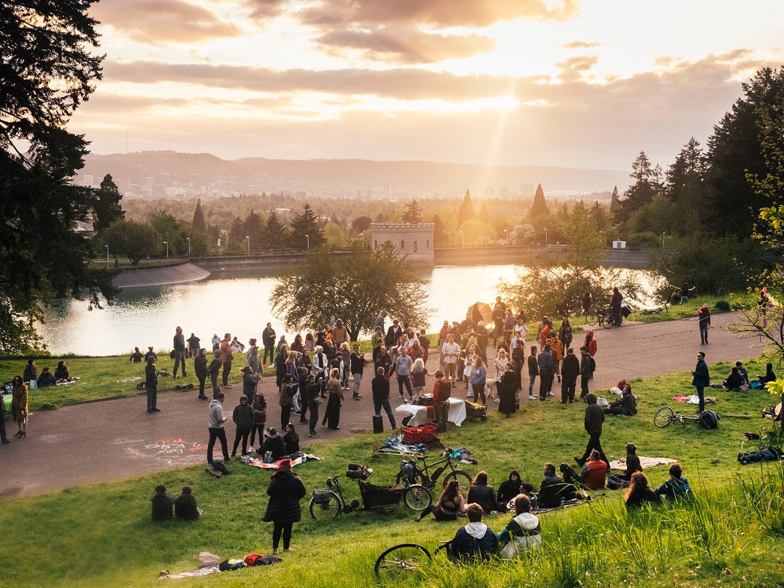 People gather in a park near a lake at sunset, sitting on the grass and standing in groups. The sun shines through clouds, casting a warm glow over the scene with trees, water, and cityscape in the background.