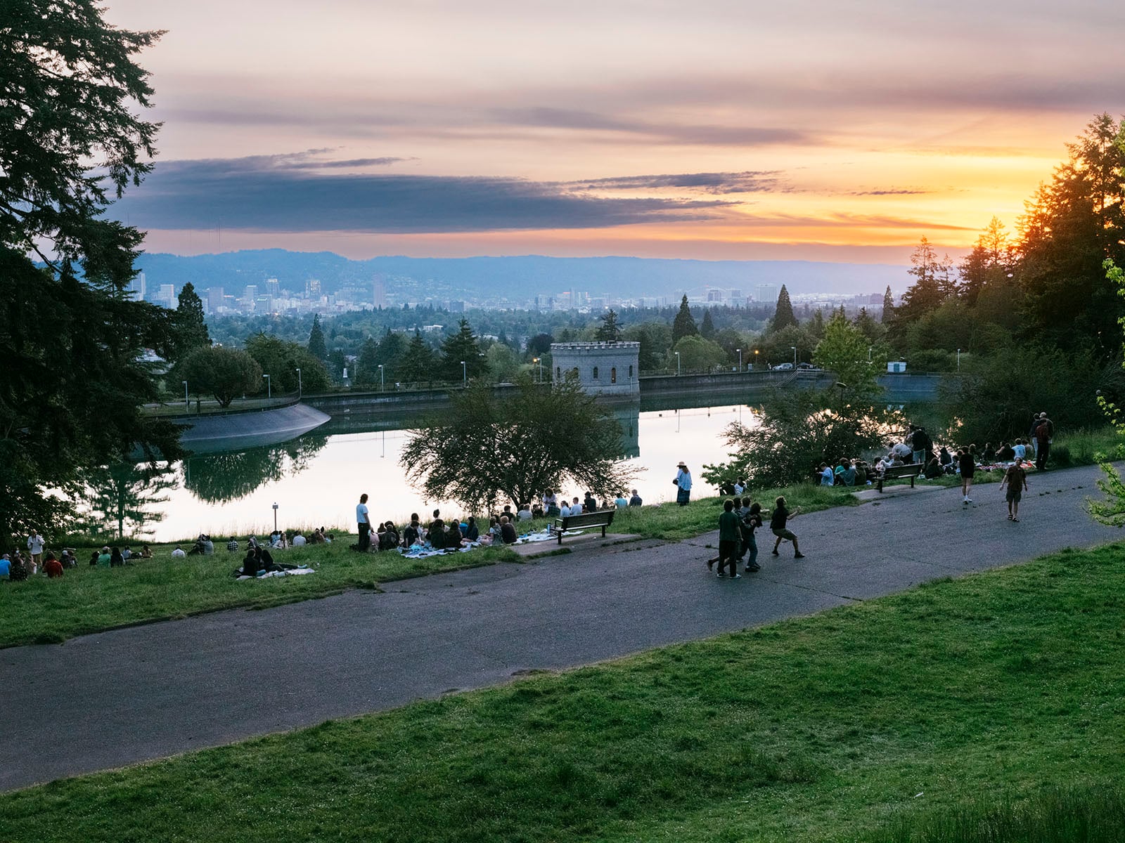 People gather on a grassy hillside overlooking a reservoir at sunset, with trees and city buildings visible in the distance under a colorful sky.