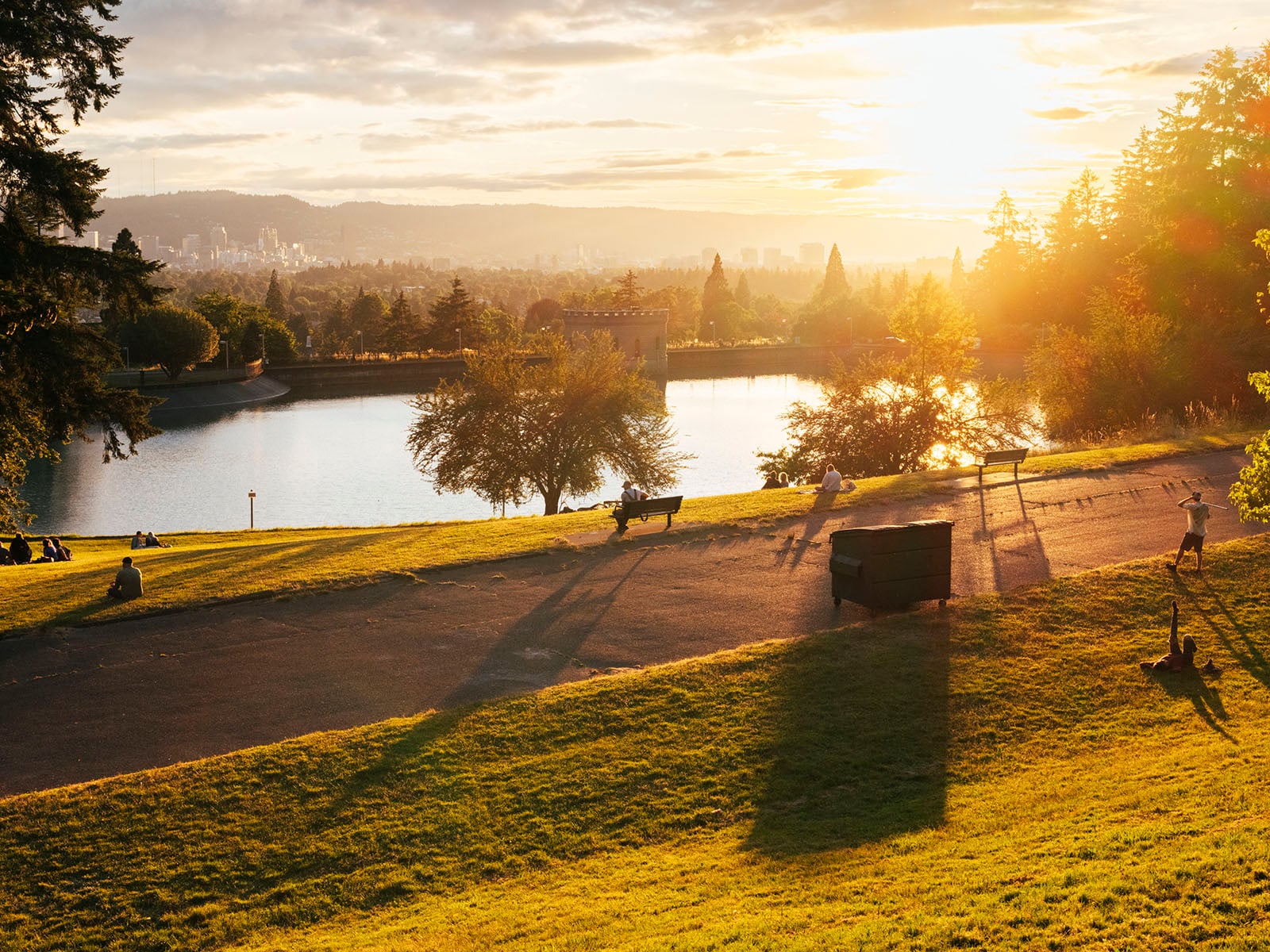 A sunlit park with a pond, trees, and benches. Long shadows stretch across the grassy hill as the sun sets, with a city skyline visible in the distant background.