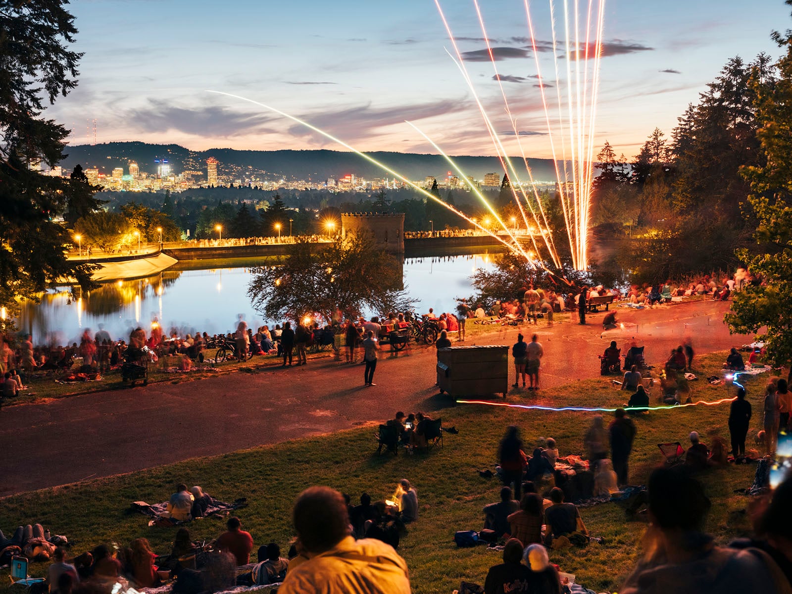 A crowd gathers on a grassy hill at dusk, watching fireworks by a riverside. City lights glow in the distance, and trees frame the scene. People sit and stand, enjoying the festive outdoor atmosphere.