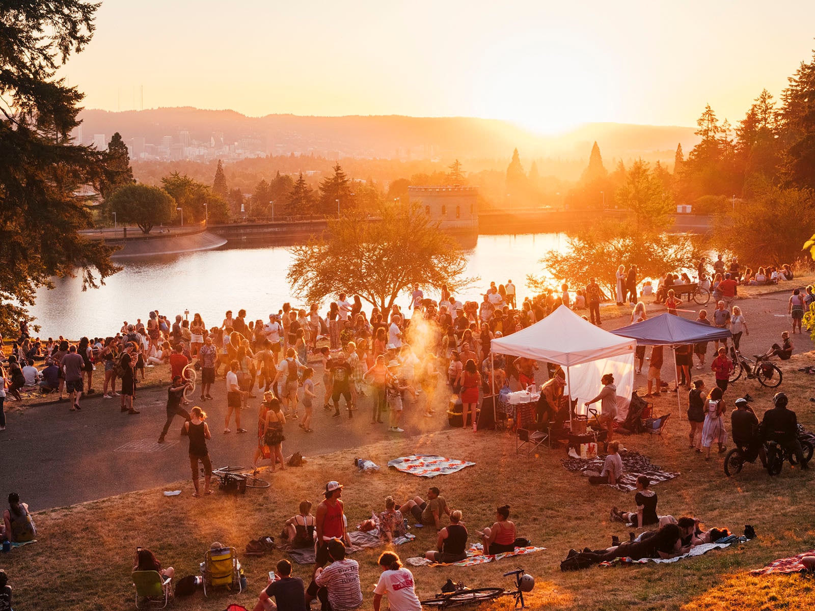 A large crowd gathers near a lake at sunset, enjoying a festival with tents, bikes, picnic blankets, and trees in the background. Warm sunlight creates a festive, relaxed atmosphere.