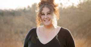 A woman with curly hair in a loose bun, wearing a black sweater and layered necklaces, stands outdoors smiling with a blurred, sunlit natural background.
