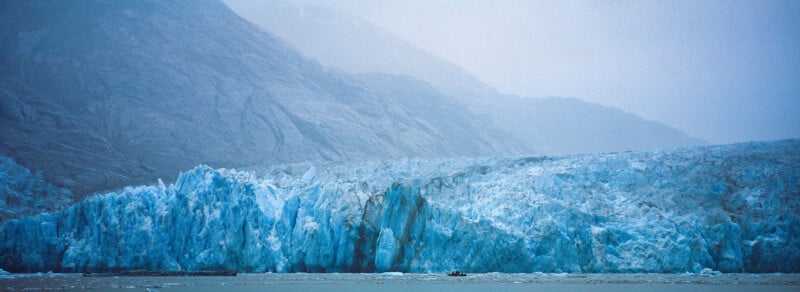 A large blue glacier meets the sea with rugged, misty mountains rising in the background. The icy landscape looks cold and dramatic, with a small boat near the glacier’s base for scale.