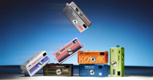 Six colorful Kodak Ektar H35 cameras are stacked and balanced creatively, with one camera hovering above the rest against a blue gradient background. The cameras are in silver, pink, blue, orange, black, and green.