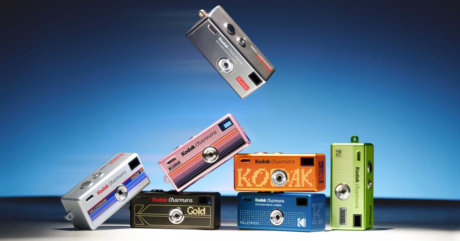 Six colorful Kodak Ektar H35 cameras are stacked and balanced creatively, with one camera hovering above the rest against a blue gradient background. The cameras are in silver, pink, blue, orange, black, and green.