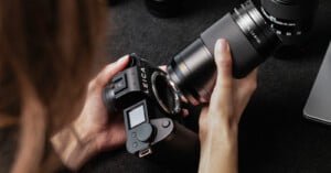A person holds a Leica camera body in one hand and a detached camera lens in the other, preparing to attach the lens. The scene is set on a dark surface with other camera equipment nearby.