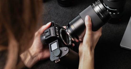 A person holds a Leica camera body in one hand and a detached camera lens in the other, preparing to attach the lens. The scene is set on a dark surface with other camera equipment nearby.