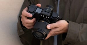 A person wearing a brown jacket holds a black Leica camera with both hands, showing the camera's controls, lens, and prominent brand name.
