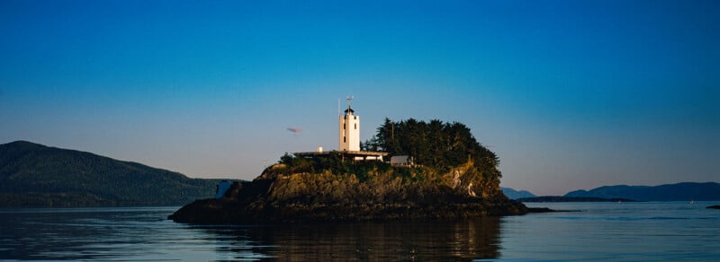 A small rocky island with a white lighthouse and trees, surrounded by calm water and distant mountains under a clear blue sky at sunset.