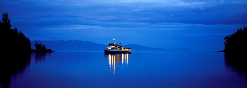 A brightly lit boat floats on calm water at dusk, reflecting its lights on the blue surface, with silhouetted trees on both sides and distant mountains under a cloudy sky.
