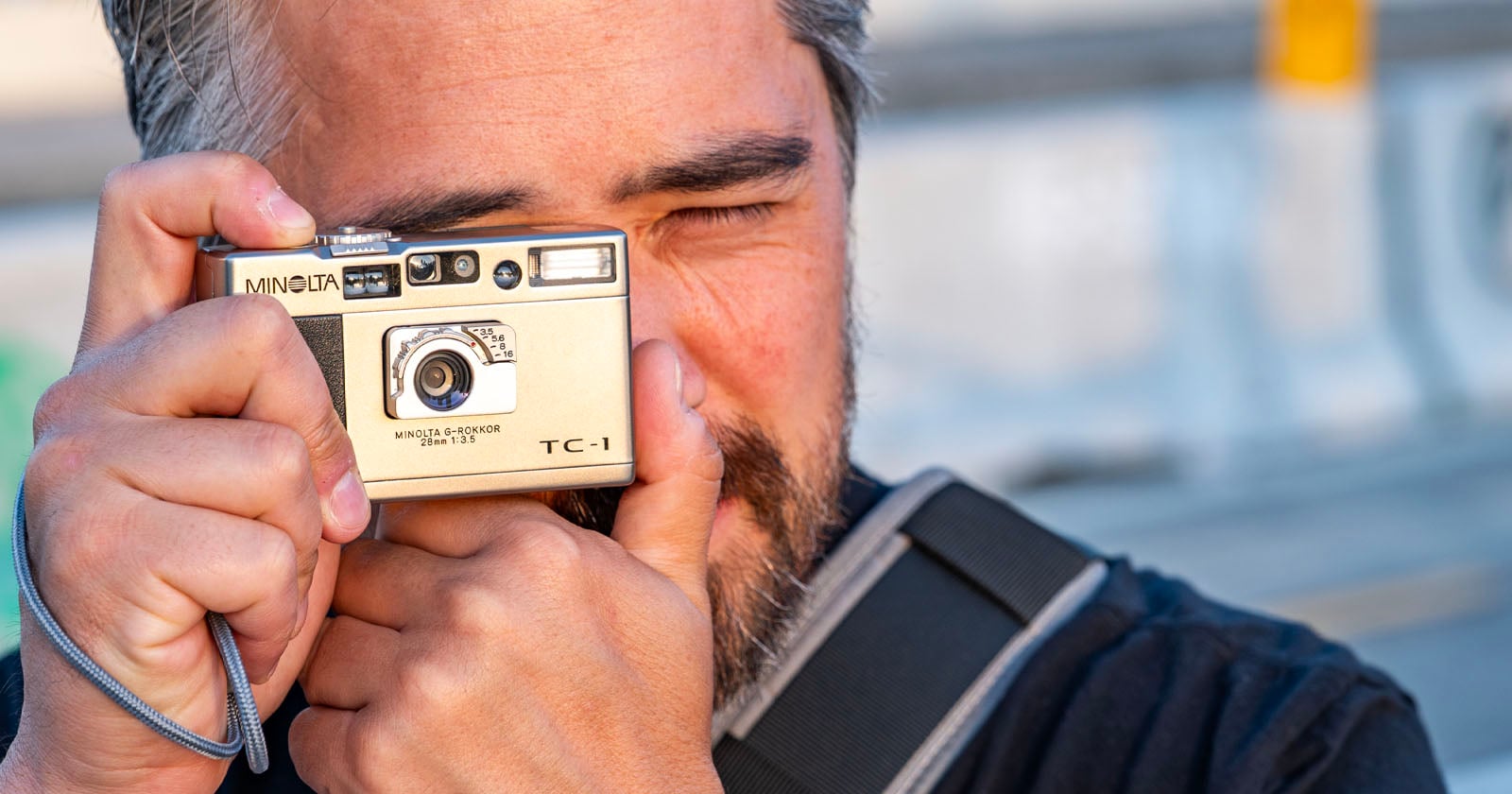 A person with facial hair holds a gold Minolta TC-1 film camera up to their face, preparing to take a photo. The background is blurred, making the camera and face the focal points.