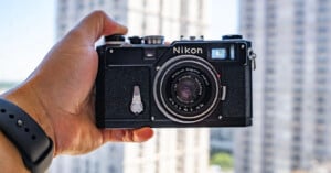 A hand holding a vintage Nikon film camera in front of a window with tall, blurry buildings in the background. The person is wearing a black wristband.