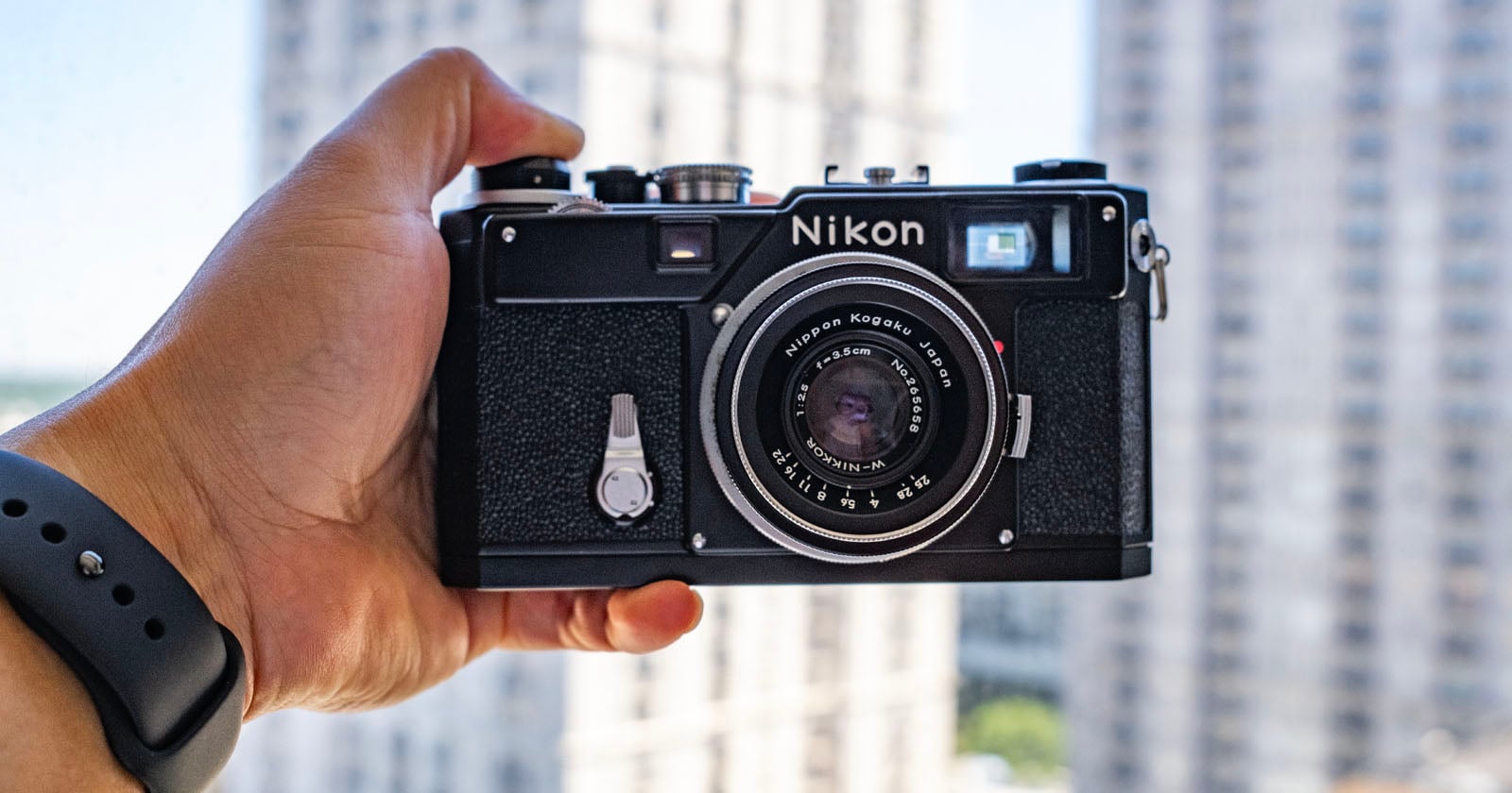 A hand holding a vintage Nikon film camera in front of a window with tall, blurry buildings in the background. The person is wearing a black wristband.