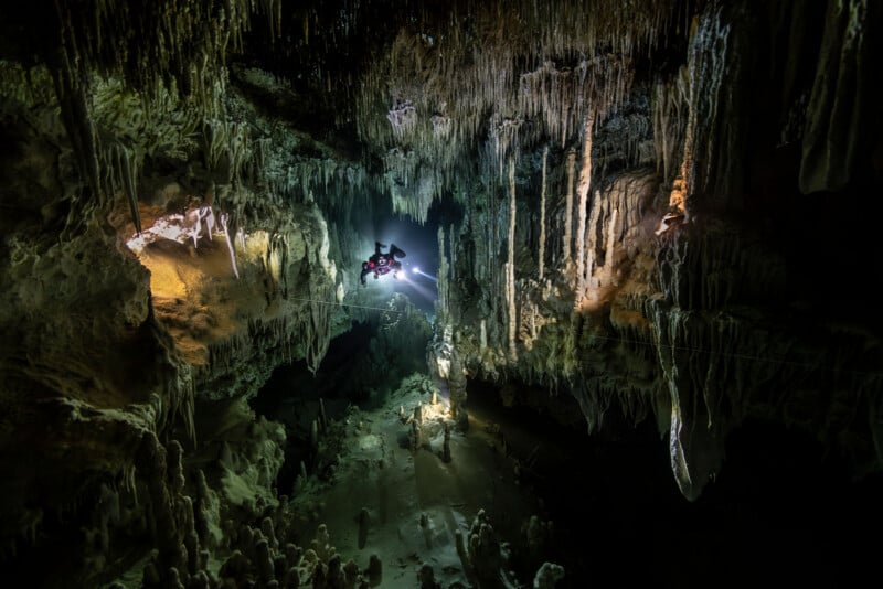 A scuba diver with a flashlight explores a large, dark cave filled with dramatic stalactites and stalagmites, illuminating parts of the rocky formations.