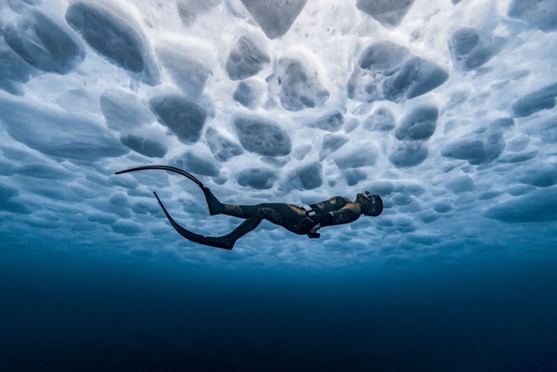 A freediver in a wetsuit and fins swims just below a thick, textured layer of ice on the water’s surface, surrounded by deep blue water.