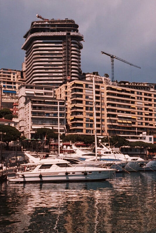 Luxury yachts docked in a marina with tall modern and older apartment buildings in the background; a construction crane is visible near the tallest building. The water reflects the boats and buildings.