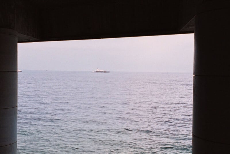 View of a calm sea with a distant yacht, framed by large pillars and the shadow of an overhanging structure at the top of the image. The sky is pale and slightly overcast.