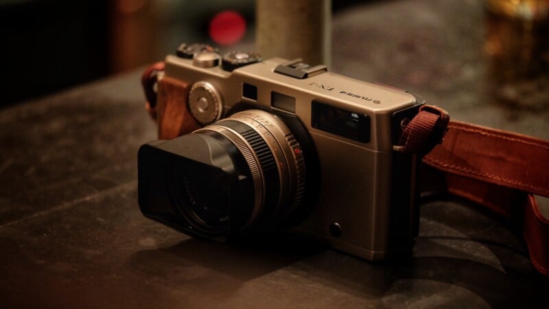A vintage-style digital camera with a brown leather strap sits on a dark surface, featuring a large lens and metal body with dials and buttons visible on top. The background is blurred.