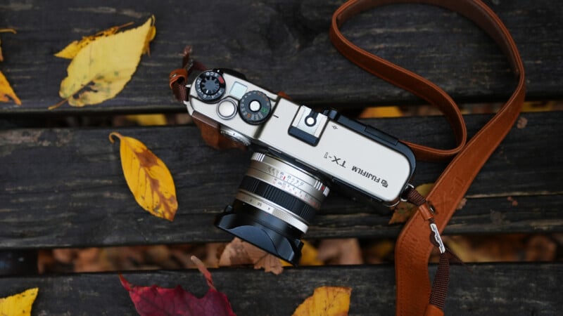 A silver Fujifilm TX-1 camera with a brown leather strap rests on a weathered wooden bench, surrounded by scattered yellow and red autumn leaves.