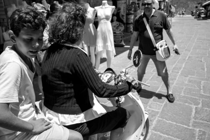 A black-and-white photo shows a boy and a woman sitting on a scooter. Mannequins in dresses stand behind them, while a man with sunglasses and a shoulder bag walks toward them on a sunlit, cobblestone street.