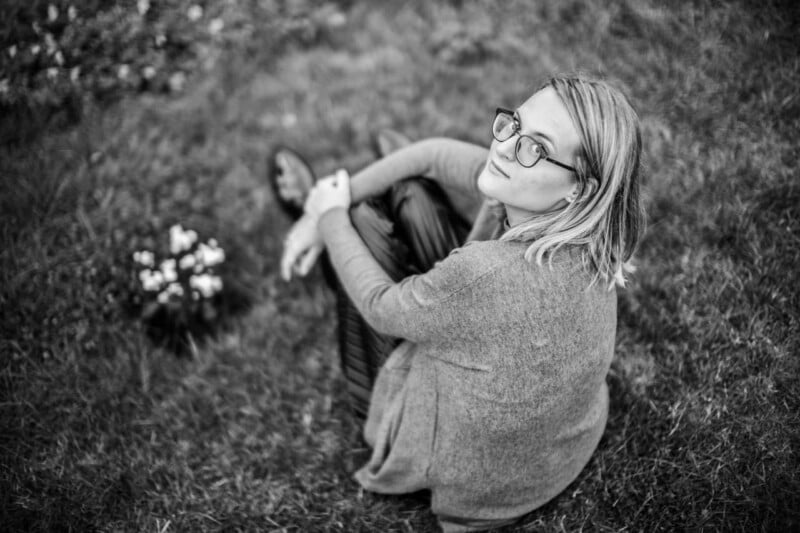 A person with glasses and shoulder-length hair sits on grass, looking up at the camera. They wear a long cardigan and are near a small bunch of flowers. The image is in black and white.