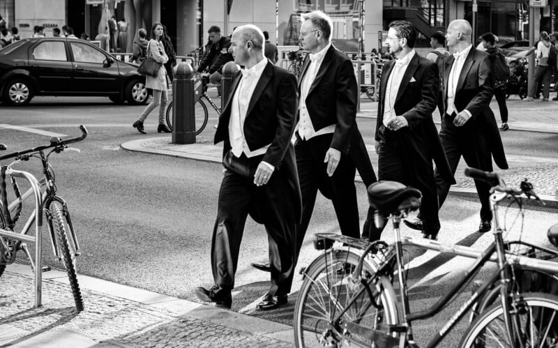 Four men in formal tailcoats and bow ties walk across a city street, passing by parked bicycles and pedestrians on the sidewalk. The scene is in black and white and set in an urban environment.