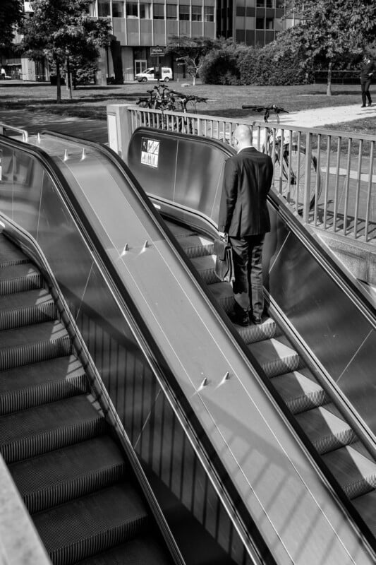 A man in a suit carries a briefcase while walking up a stationary escalator outdoors, next to a working escalator. The scene is in black and white, with buildings and trees in the background.