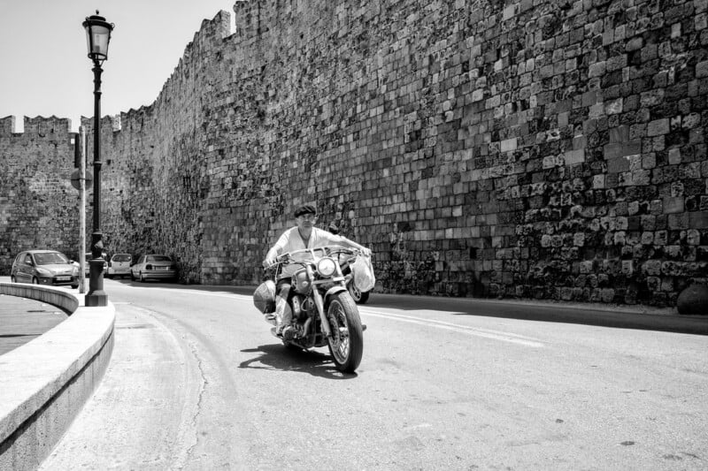 A man rides a motorcycle along a curving road next to a large, ancient stone wall. Parked cars are visible along the wall, and a streetlamp stands on the left. The scene is in black and white.