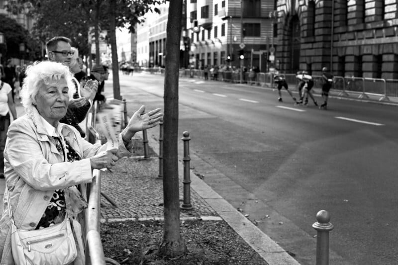 Elderly woman and man stand on a city sidewalk, clapping as rollerbladers race down an empty street lined with buildings and barriers. The scene is in black and white.
