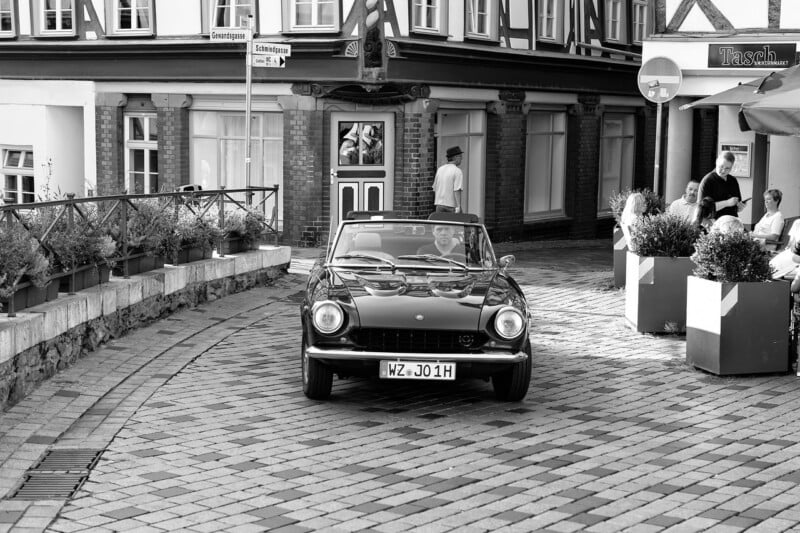 A classic convertible car drives on a cobblestone street in front of historic buildings, while people sit and relax at outdoor tables nearby. The image is in black and white.