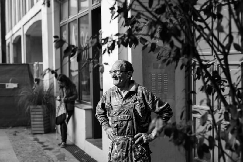 A man wearing paint-splattered overalls stands with his hands on his hips outside a building, looking to the side. Another person stands in the background near a window, partially obscured by plants. The image is in black and white.