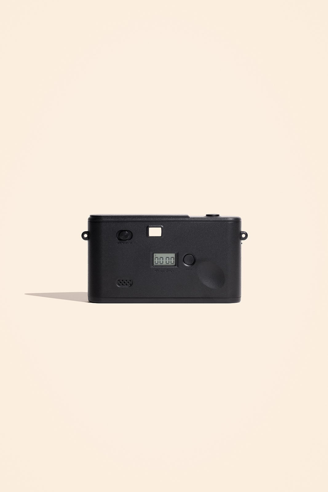A black digital camera is positioned upright on a light beige background, shown from the back with its screen and buttons visible. The camera casts a shadow to the left.