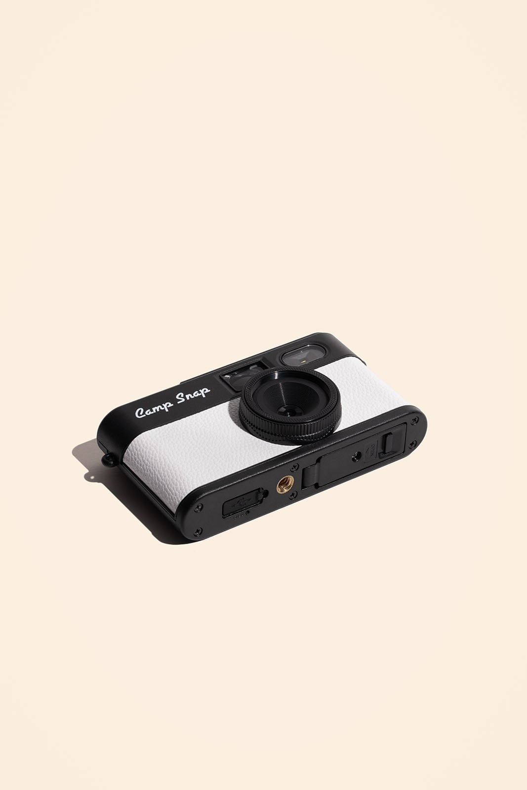 A retro-style black and white digital camera with "Camp Snap" written on it, positioned on a light beige background with a shadow cast to the left.