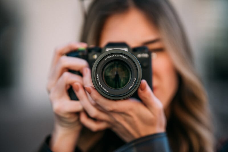 A person with long hair holds a camera up to their face, focusing the lens directly at the viewer. The background is blurred, drawing attention to the camera and hands in the foreground.
