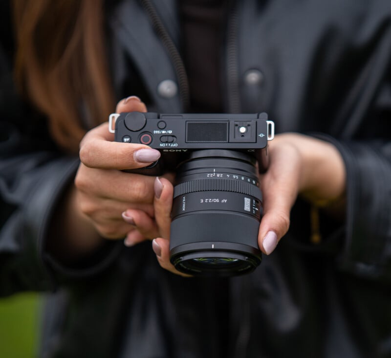 A person wearing a black leather jacket holds a digital camera with both hands, focusing on the camera's lens and buttons. The background is blurred.