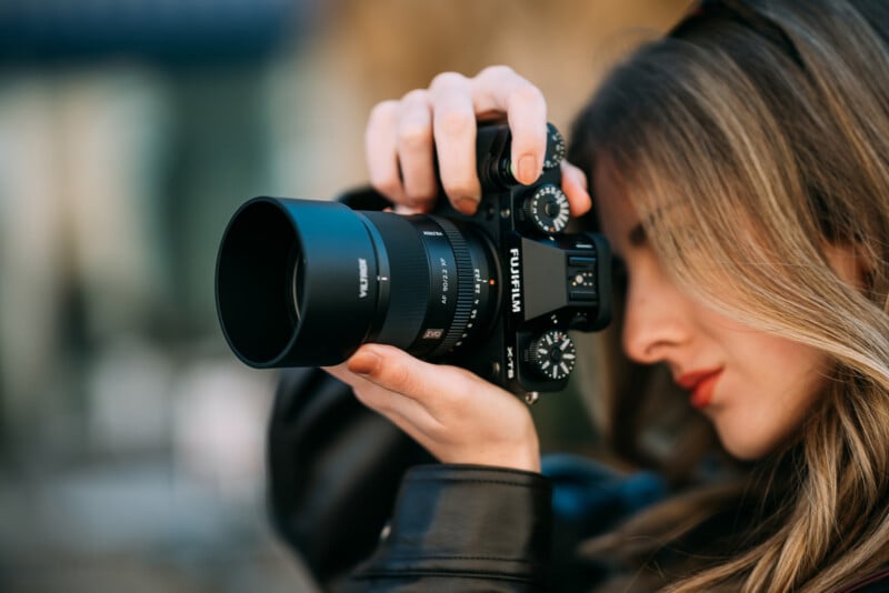 A woman with long hair holds a Fujifilm camera up to her eye, focusing intently as she prepares to take a photograph outdoors.