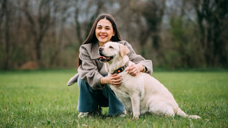 A smiling woman in a gray coat crouches on grass, holding a happy yellow Labrador retriever. Trees and greenery are blurred in the background, suggesting a park or outdoor setting.