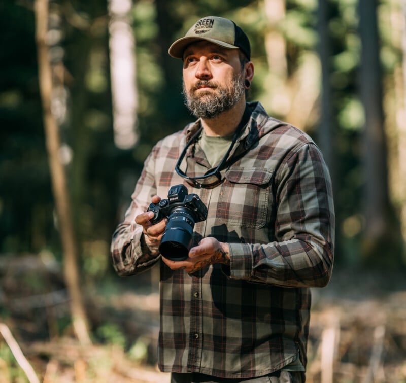 A bearded man wearing a plaid shirt and cap stands in a sunlit forest, holding a camera and looking upward thoughtfully.