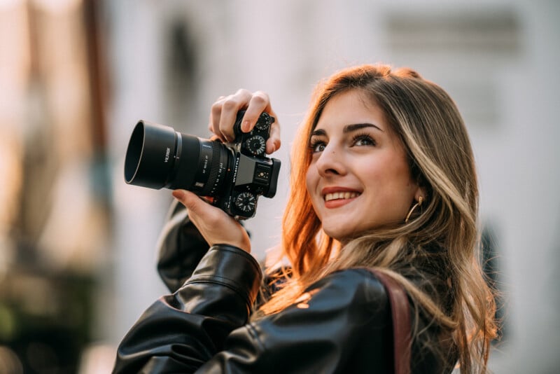 A young woman with long, light brown hair and a leather jacket smiles while holding a camera up to her face, getting ready to take a photo outdoors.