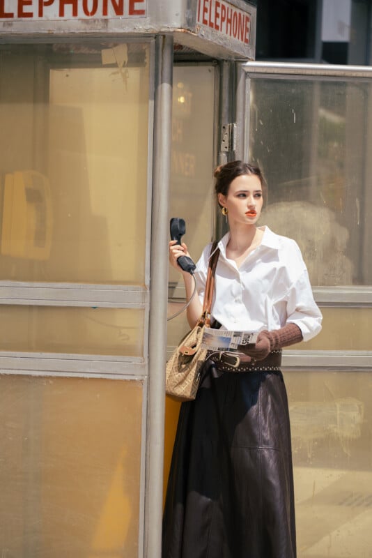 A stylish woman in a white blouse and long dark skirt stands in a vintage phone booth, holding a telephone receiver and looking outside while holding a newspaper and a tan handbag.