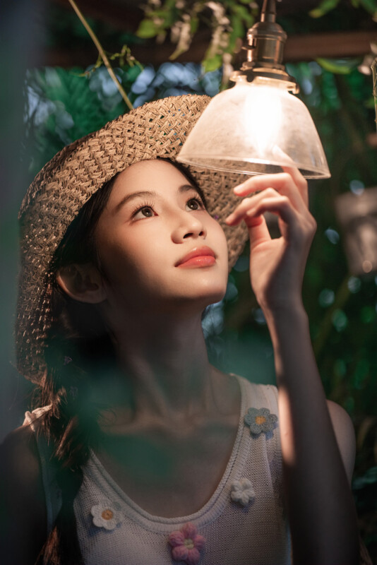 A young woman wearing a straw hat and a sleeveless top with flower decorations gazes up and gently touches a hanging light bulb, surrounded by greenery. Soft, warm light illuminates her face.