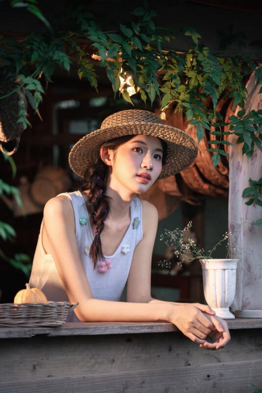 A young woman with a braid, wearing a straw hat and sleeveless top, leans on a wooden counter surrounded by greenery. Soft evening light illuminates her face, and a white cup with flowers sits in front of her.