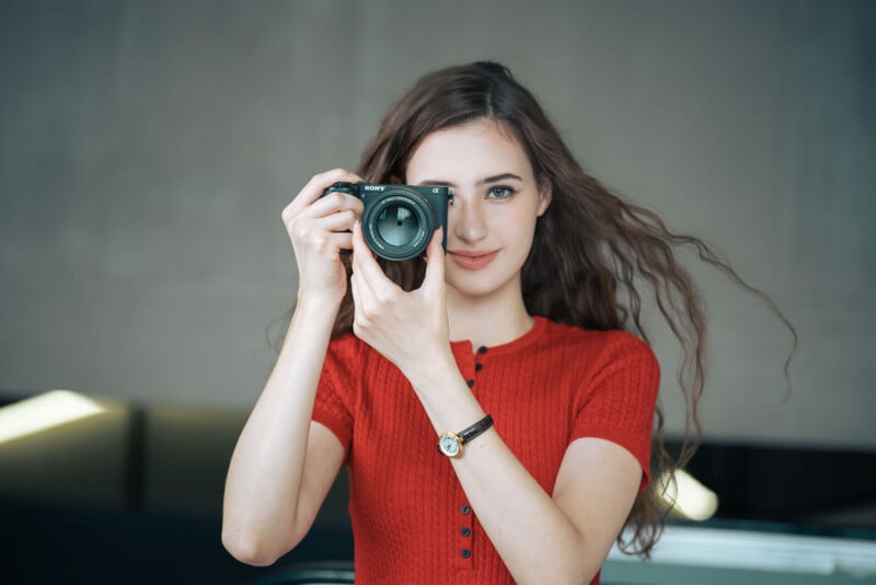 A woman with long brown hair, wearing a red short-sleeve top and a wristwatch, holds a camera up to her eye and smiles gently at the viewer. The background is softly blurred.