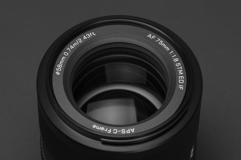 Close-up view of a camera lens with details visible, including "AF 75mm 1:1.8 STM ED IF" and "APS-C Frame" printed on the rim, set against a dark background.