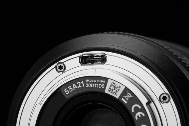 Close-up of a camera lens mount showing metallic contacts, screws, a barcode, text “Made in China,” and certification symbols, all against a black background.