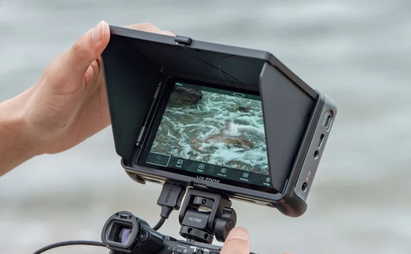 A person holds a camera monitor with a sunshade, displaying a close-up video of moving water or waves, while filming near a body of water.
