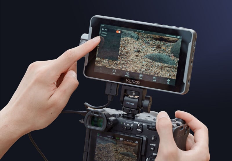 A person adjusts settings on a Viltrox external camera monitor mounted on a digital camera, with the monitor displaying a close-up image of pebbles and rocks.