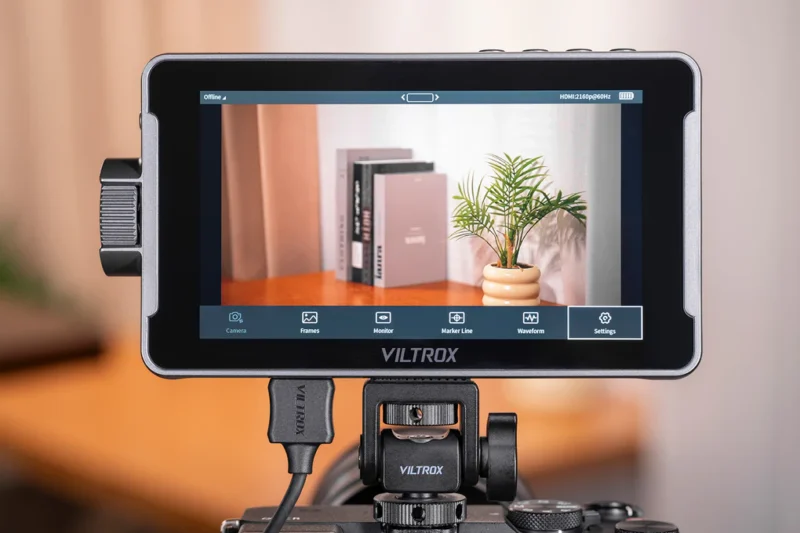 A Viltrox camera monitor displays a focused image of a small table with books and a potted plant on its screen. The monitor interface shows several camera settings and options at the bottom.