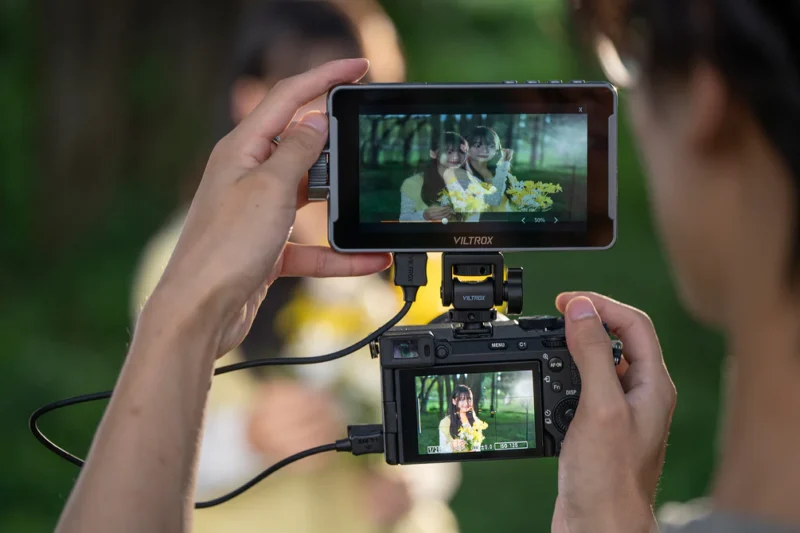 A person holds a camera and an external monitor, both displaying a clear image of a woman holding a bouquet of yellow flowers outdoors, while the actual woman appears blurred in the background.