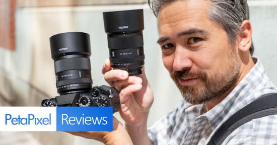 A man holds a camera with a large lens in one hand and a second lens in the other, standing outdoors. The image includes “PetaPixel Reviews” text in the lower left corner.